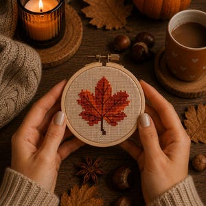 May include: A circular wooden embroidery hoop containing a cross-stitch design of a single maple leaf in shades of orange, red, and brown on a light beige fabric background. The hoop features a metal tightening screw at the top centre.