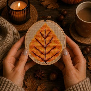 May include: A circular wooden embroidery hoop containing a cross-stitch pattern of a single autumn leaf. The leaf design features a gradient of yellow, orange, and red thread colours with a dark brown central vein and lateral veins on beige fabric.