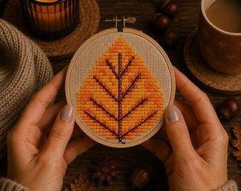 May include: A circular wooden embroidery hoop containing a cross-stitch pattern of a single autumn leaf. The leaf design features a gradient of yellow, orange, and red thread colors with a dark brown central vein and lateral veins on beige fabric.