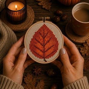 May include: A circular wooden embroidery hoop containing a cross-stitch pattern of a single autumn leaf. The leaf is rendered in shades of orange, red, and brown thread on a light beige fabric background. The hoop features a metal tightening screw.