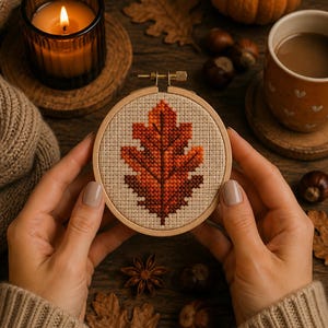 May include: A circular wooden embroidery hoop containing a cross-stitch design of an autumn oak leaf. The leaf features a gradient of orange, red, and brown thread colours stitched onto a light beige fabric background with a visible grid texture.