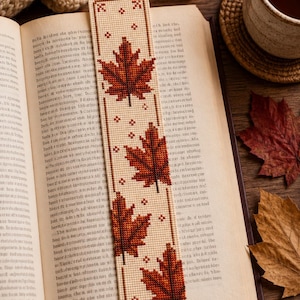 May include: A rectangular bookmark featuring a cross-stitch pattern of three dark red and brown maple leaves on a cream-coloured fabric background, accented with small brown cross-stitch details and a decorative border on the top and bottom edges.