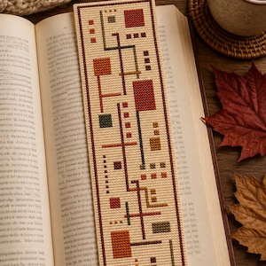 May include: A rectangular bookmark featuring a cross-stitch pattern of geometric shapes in shades of rust, olive green, and dark brown on a cream-colored fabric background, with a thin dark brown border framing the entire design.