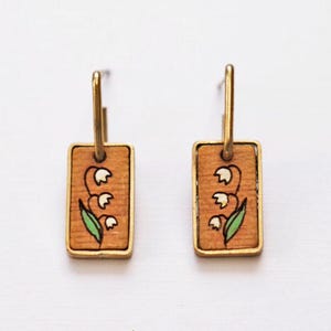 May include: A pair of gold hoop earrings with rectangular wooden pendants. Each pendant features a hand-painted lily design with white flowers and green leaves. The word "MAY" is above the earrings, and the word "Lily" is below, with the phrase "sweetness, joy, motherhood".