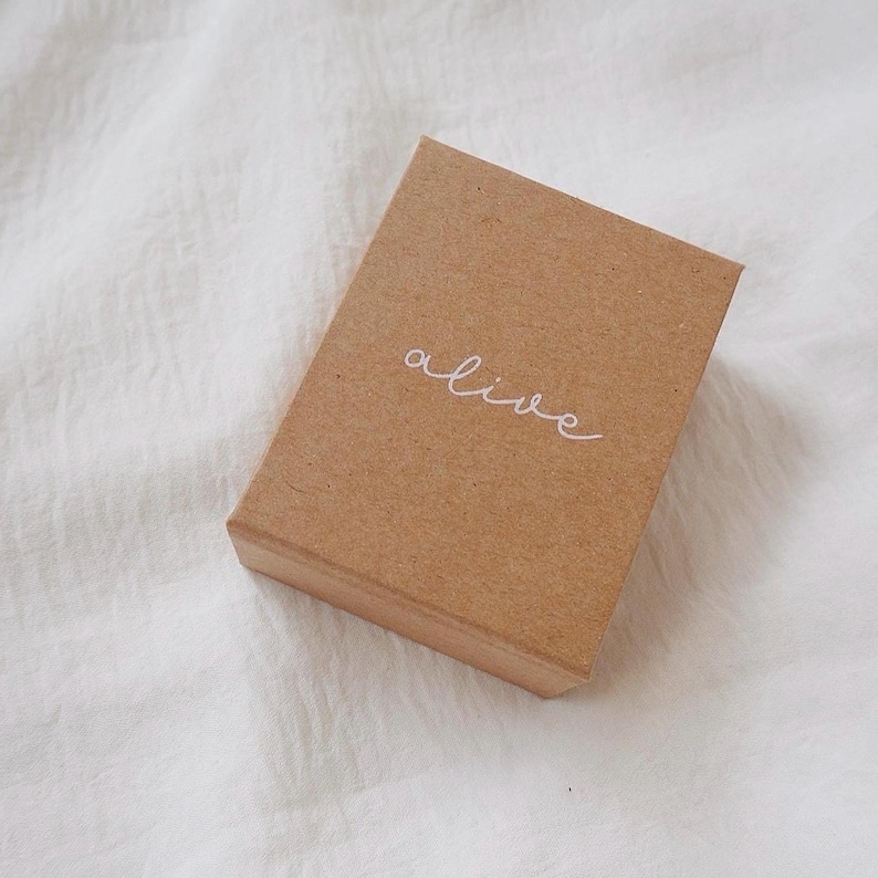 May include: A small, rectangular, brown cardboard box with the word "alive" written in white cursive script on the top. The box is set against a white, textured background.