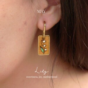May include: Gold-tone hoop earring with a rectangular pendant. The pendant features a hand-painted lily of the valley design on a wood-grain background. The word "MAY" is at the top, and "Lily" is written below the earring.