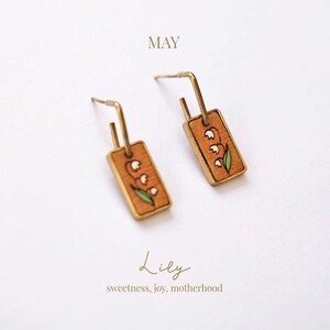 May include: A pair of gold-toned earrings with rectangular wooden pendants. Each pendant features a hand-painted lily design with white flowers and green leaves. The word "MAY" is above the earrings, and the word "Lily" with the text "sweetness, joy, motherhood" is below.