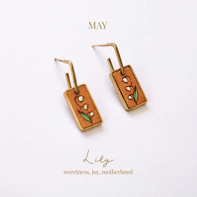 May include: A pair of gold-toned earrings with rectangular wooden pendants. Each pendant features a hand-painted lily design with white flowers and green leaves. The word "MAY" is above the earrings, and the word "Lily" with the text "sweetness, joy, motherhood" is below.