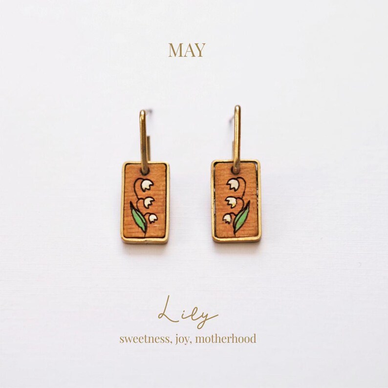 May include: A pair of gold hoop earrings with rectangular wooden pendants. Each pendant features a hand-painted lily design with white flowers and green leaves. The word "MAY" is above the earrings, and the word "Lily" is below, with the phrase "sweetness, joy, motherhood".