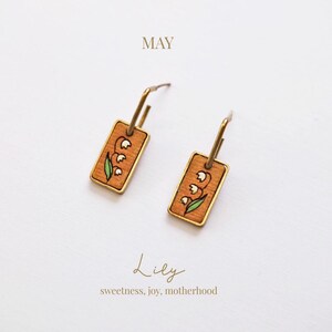 May include: A pair of gold-tone earrings with rectangular wooden pendants. Each pendant features a hand-painted lily of the valley design with white flowers and green leaves. The word "MAY" is at the top, and "Lily" and "sweetness, joy, motherhood" are at the bottom.