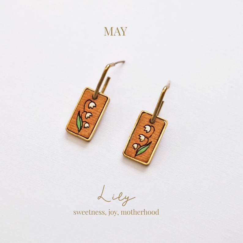 May include: A pair of gold-tone earrings with rectangular wooden pendants. Each pendant features a hand-painted lily of the valley design with white flowers and green leaves. The word "MAY" is at the top, and "Lily" and "sweetness, joy, motherhood" are at the bottom.