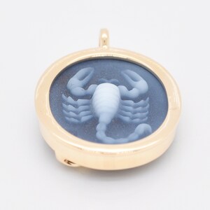 Pode incluir: Pingente dourado com um centro circular azul escuro com um desenho de escorpi&atilde;o branco. O pingente tem uma argola na parte superior para uma corrente. O escorpi&atilde;o &eacute; o ponto focal, com suas garras detalhadas e corpo segmentado.