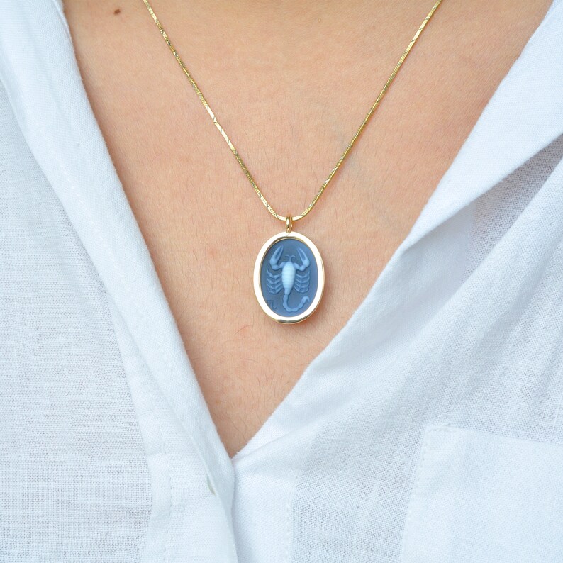 Pode incluir: Colar dourado com um pingente oval com um design de escorpi&atilde;o azul claro. O pingente, emoldurado em ouro, pende de uma delicada corrente de ouro. O colar &eacute; exibido contra uma camisa branca.