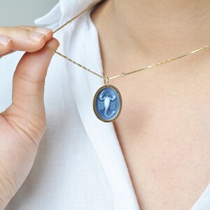 Pode incluir: Um colar dourado com um pingente oval. O pingente apresenta uma camafeu azul com um desenho de escorpi&atilde;o branco. O colar &eacute; segurado por uma pessoa vestindo uma camisa branca.