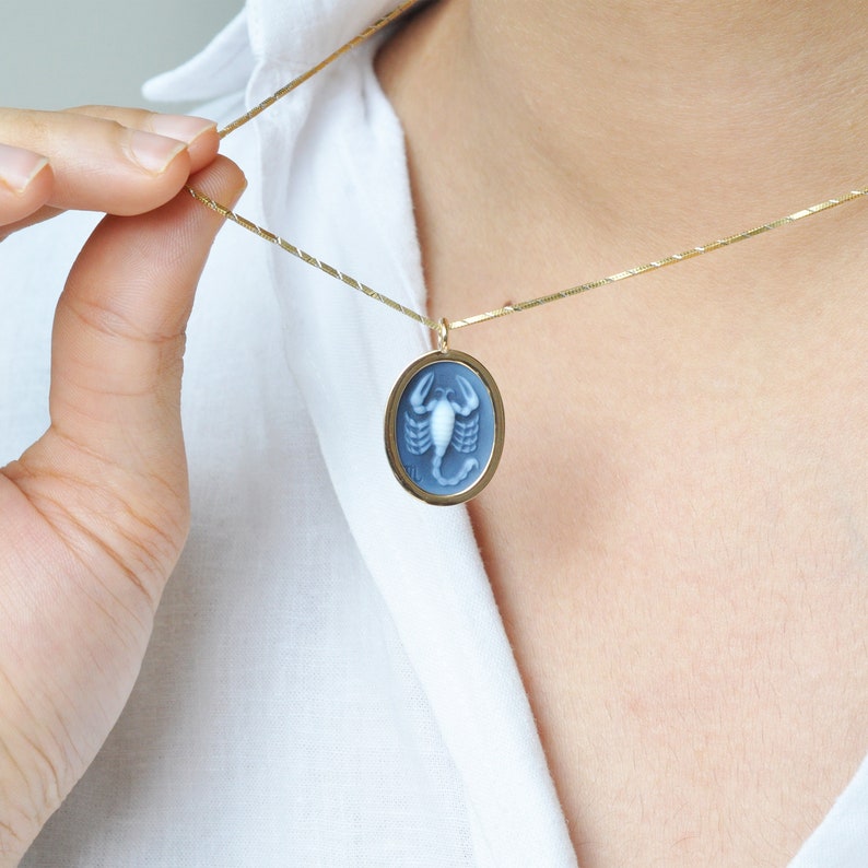 Pode incluir: Um colar dourado com um pingente oval. O pingente apresenta uma camafeu azul com um desenho de escorpi&atilde;o branco. O colar &eacute; segurado por uma pessoa vestindo uma camisa branca.