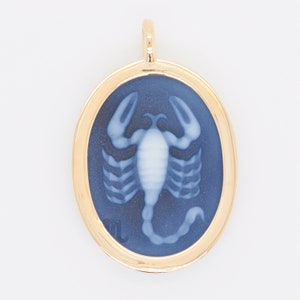 Pode incluir: Pingente dourado com um centro oval azul escuro com um desenho de escorpi&atilde;o branco. O pingente tem uma moldura dourada e uma pequena argola na parte superior para uma corrente. O escorpi&atilde;o &eacute; um s&iacute;mbolo do zod&iacute;aco.