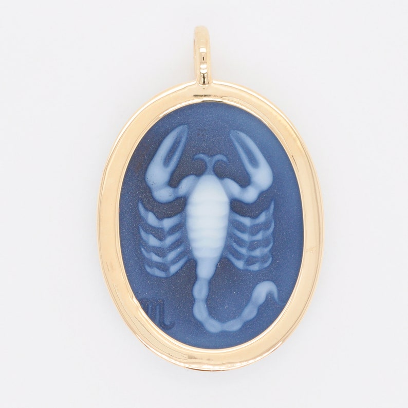 Pode incluir: Pingente dourado com um centro oval azul escuro com um desenho de escorpi&atilde;o branco. O pingente tem uma moldura dourada e uma pequena argola na parte superior para uma corrente. O escorpi&atilde;o &eacute; um s&iacute;mbolo do zod&iacute;aco.