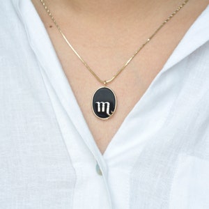 Pode incluir: Um colar de corrente dourada com um pingente oval preto. O pingente apresenta uma borda dourada e a letra branca "m", representando o signo do zod&iacute;aco Escorpi&atilde;o. O colar &eacute; usado sobre uma camisa branca.