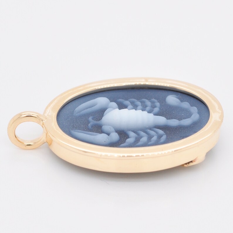 Pode incluir: Pingente oval dourado com um design detalhado de escorpi&atilde;o em tons de branco e azul. O pingente tem uma argola para corrente e um fecho na parte de tr&aacute;s. O escorpi&atilde;o &eacute; o ponto focal desta joia.