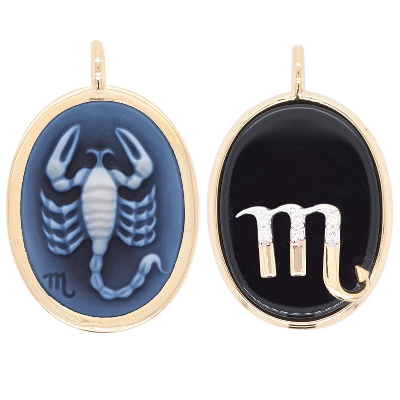 Pode incluir: Dois pingentes ovais com moldura dourada. Um apresenta um desenho de escorpi&atilde;o azul claro sobre um fundo azul mais escuro. O outro pingente tem um fundo preto com o s&iacute;mbolo de Escorpi&atilde;o, acentuado com pequenas pedras brancas.