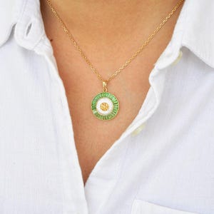 May include: Gold-toned necklace with a circular pendant. The pendant features a white center with a small gold-colored flower design, surrounded by a ring of green and white stones. The necklace is worn over a white button-down shirt.
