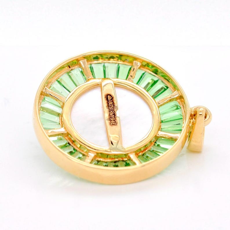 May include: A gold-toned pendant with a central white circular design, surrounded by vibrant green rectangular gemstones. The pendant has a clasp and a small bar with text. The gemstones are arranged around the white center.