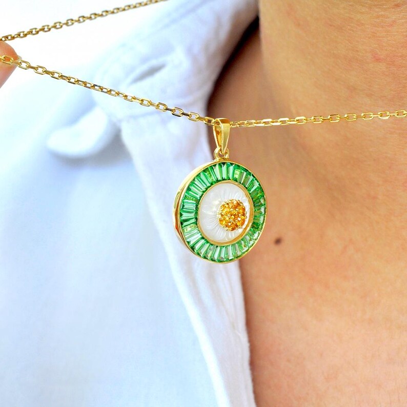 May include: A gold necklace featuring a round pendant with a white center, surrounded by green and gold accents. The pendant has a floral design with a gold chain. The necklace is worn against a white shirt.