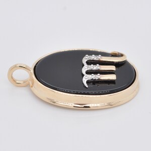 Pode incluir: Pingente dourado com um centro oval preto. O pingente apresenta a letra "E" em ouro e prata, adornada com pequenas pedras brilhantes. O pingente tem uma argola para uma corrente.