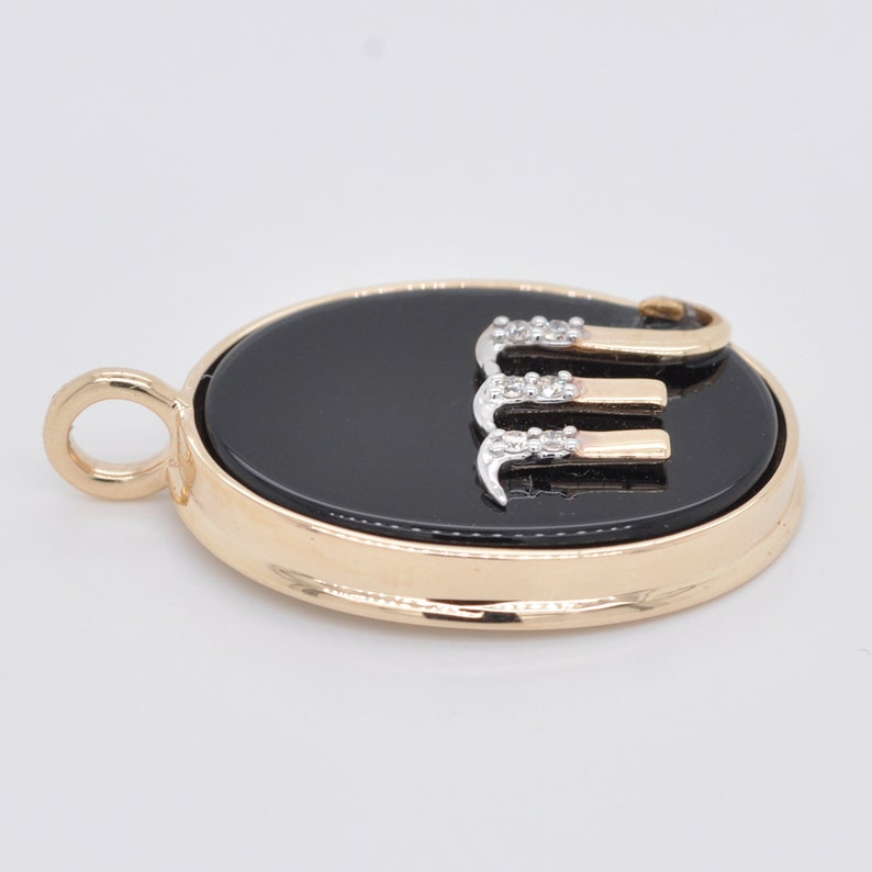 Pode incluir: Pingente dourado com um centro oval preto. O pingente apresenta a letra "E" em ouro e prata, adornada com pequenas pedras brilhantes. O pingente tem uma argola para uma corrente.