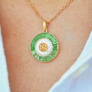 May include: A gold-toned necklace with a circular pendant. The pendant features a white center with a cluster of small, gold-colored stones, surrounded by a ring of green baguette-cut stones. The outer edge is gold, and the chain is also gold.