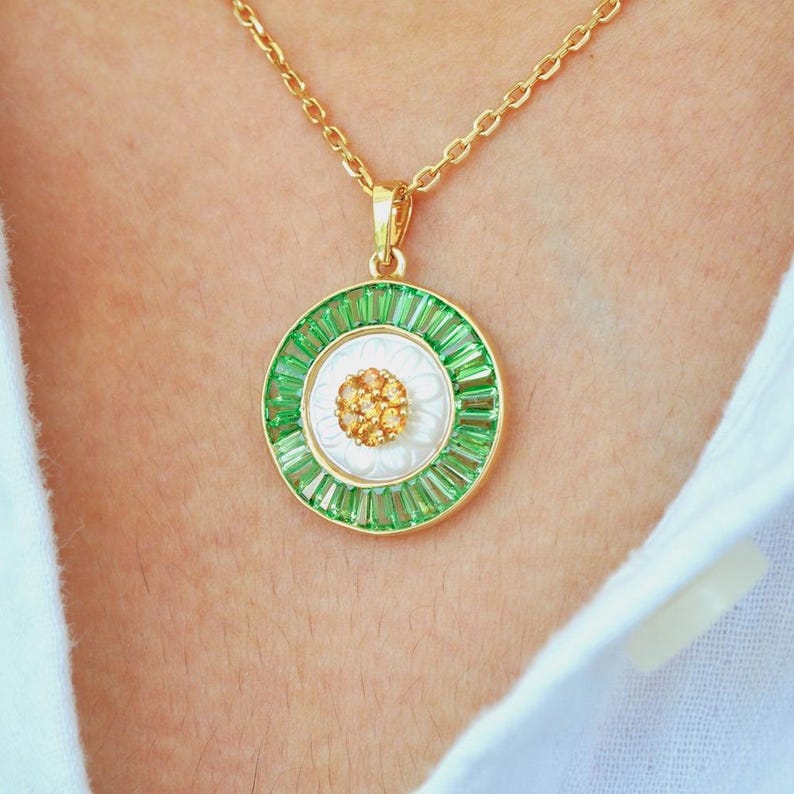May include: A gold-toned necklace with a circular pendant. The pendant features a white center with a cluster of small, gold-colored stones, surrounded by a ring of green baguette-cut stones. The outer edge is gold, and the chain is also gold.