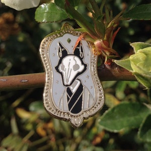 May include: Gold-toned enamel pin featuring a white skull with black eyes and a black and white patterned kimono. The pin is in the shape of a mirror with a gold frame and a diamond-shaped design at the top.