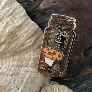 May include: Gold-tone enamel pin featuring a mushroom inside a glass jar with a label that reads "J.J. Radish Co. Est. 12"