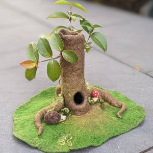 May include: A felt miniature tree with a hollow trunk, green leaves, and a small mushroom. The tree is sitting on a green felt base with a brown felt hedgehog.