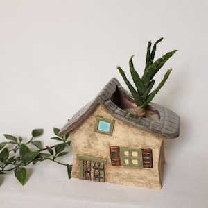 Könnte beinhalten: Ein hausförmiger Keramik-Blumentopf mit grauem Dach, beigefarbenen Wänden und grünen und braunen Akzenten. Ein kleines blaues Fenster sowie eine grüne Tür und Fensterläden sind sichtbar. Eine grüne Pflanze wächst aus dem Dach.