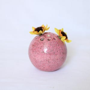 Könnte beinhalten: Eine kugelförmige, rosa Keramik-Blumenfrosch-Vase mit gesprenkelter Textur. Die Vase hat mehrere Löcher oben und hält zwei gelbe und braune Sonnenblumen. Die Vase ist ein dekorativer Artikel für Blumenarrangements.