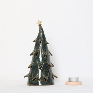 Könnte beinhalten: Ein Kerzenhalter in Form eines Weihnachtsbaums aus Keramik mit tealfarbener Glasur und goldenen Akzenten. Der Baum hat ausgeschnittene Formen und einen goldenen Stern oben. Ein kleiner, runder Kerzenhalter befindet sich neben dem Baum. Der Baum ist etwa 18 cm hoch.