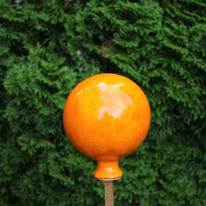 Könnte beinhalten: Ein glänzendes, kugelförmiges Gartenornament aus orangefarbener Keramik, das auf einem dünnen, hellbraunen Holzstab montiert ist. Die Oberfläche der Kugel ist glatt und reflektierend mit einer leuchtenden, gleichmäßigen Glasur.