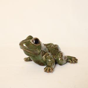 Könnte beinhalten: Eine Froschfigur aus Keramik mit einer glänzenden, gesprenkelten dunkelgrünen Glasur. Die Skulptur hat einen strukturierten Rücken, einen glatten Kopf und große, dunkle, abgerundete Augen. Der Frosch sitzt in einer ruhenden Position auf dem Boden.