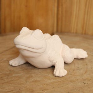 Könnte beinhalten: Eine matte, cremeweiße Froschfigur aus Keramik in ruhender Position. Die Skulptur hat einen glatten Kopf und Rücken mit strukturierten, noppigen Hautdetails an den Beinen und Seiten. Der Frosch hat hervorstehende Augen und Schwimmfüße.