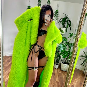 May include: A neon green faux fur coat with a long, flowing design. The coat is paired with black lingerie and boots. The image is taken in front of a mirror, with plants visible in the background.