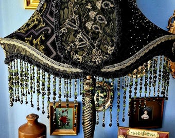 Può includere: Un'antica lampada ornata con un paralume nero decorato con pizzo, ricami dorati e frange di perline. La lampada è su un tavolo di legno con oggetti vintage, tra cui foto incorniciate e una bottiglia marrone.