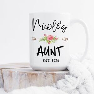 May include: White ceramic mug with black text that reads "Nicole's Aunt Est. 2020". A floral design with pink flowers and green leaves is above the text with a brown arrow pointing to the left and right.