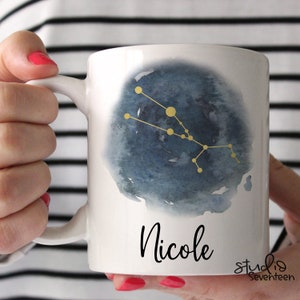 May include: White ceramic mug with a watercolor design of the Taurus constellation in gold on a blue background. The name "Nicole" is written in black cursive below the design. The mug is held by a hand.