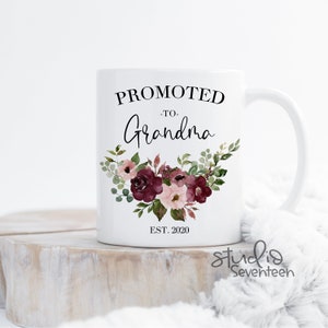 May include: White ceramic mug with the text "Promoted to Grandma Est. 2020" in black lettering. The mug features a floral design with burgundy and pink flowers and green leaves.