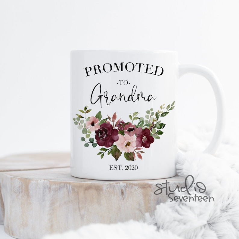 May include: White ceramic mug with the text "Promoted to Grandma Est. 2020" in black lettering. The mug features a floral design with burgundy and pink flowers and green leaves.