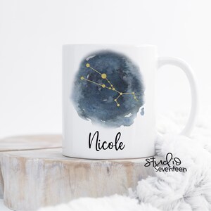 May include: White ceramic mug with a blue watercolor design featuring the Taurus constellation in gold. The mug has the name "Nicole" printed on the front.
