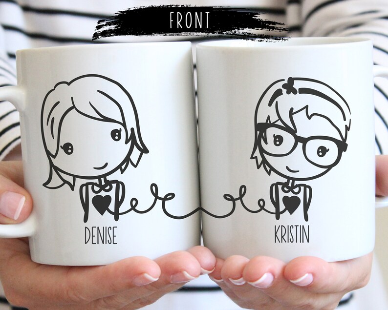 May include: Two white ceramic mugs with black line drawings of women wearing glasses and holding hearts. The mugs are connected by a line and the text "Denise" and "Kristin" is printed below the drawings.