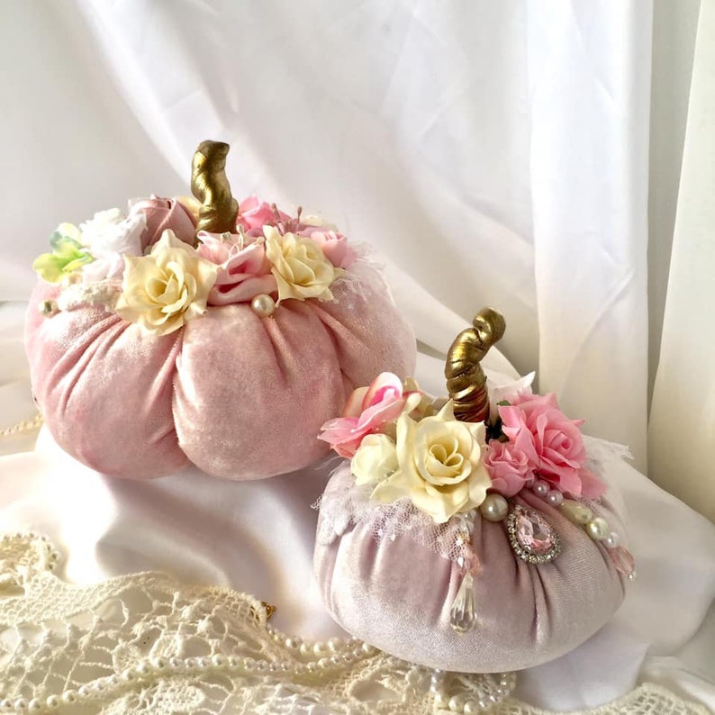May include: Two pink velvet pumpkins decorated with white and pink flowers, gold accents, and pearl-like embellishments.