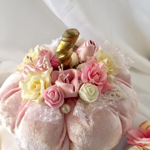 May include: A pink velvet pumpkin decorated with white lace, pink and white roses, and a gold stem.